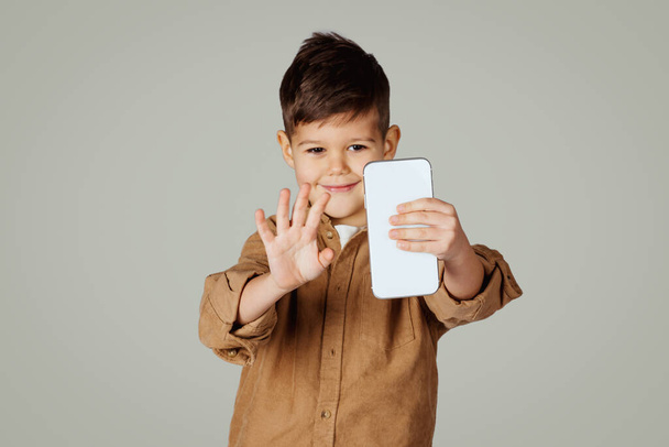 明るい白人6歳の男の子がスマートフォンに手を振って、灰色のスタジオの背景に隔離されたビデオ通話を持っています。デバイスとの通信のためのアプリ,ライフスタイルと遠隔授業 - 写真・画像