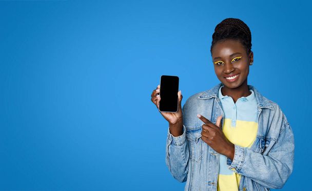 Весела мила красива молода чорна жінка випадково вказувала на мобільний телефон з порожнім екраном в руці, показуючи гарну онлайн-пропозицію, макет, ізольована на синьому фоні студії, копіювати простір
 - Фото, зображення