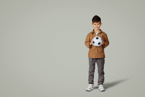 Lächelnde kaukasische 6 Jahre alten kleinen Jungen in lässig mit Fußball, genießen Spiel, isoliert auf grauem Studiohintergrund. Zukunft mit Sport, Hobby und Spiel, kindlichen Emotionen - Foto, Bild