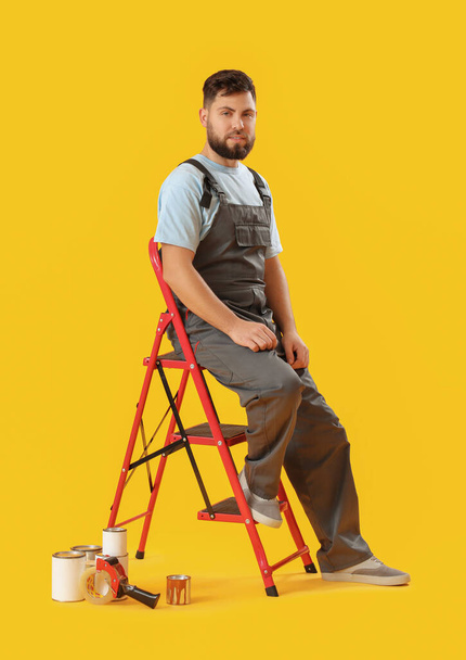Male worker with ladder and cans of paint on yellow background - Foto, Imagen