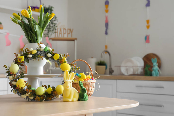 Florero con tulipanes, corona de Pascua, conejos y canasta de huevos sobre mesa en cocina - Foto, Imagen