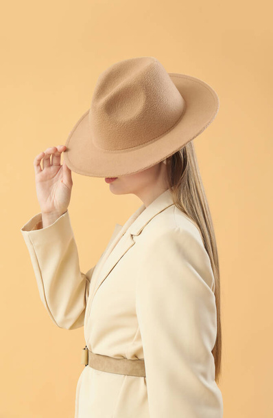 Fashionable young woman in stylish jacket and hat on beige background - 写真・画像