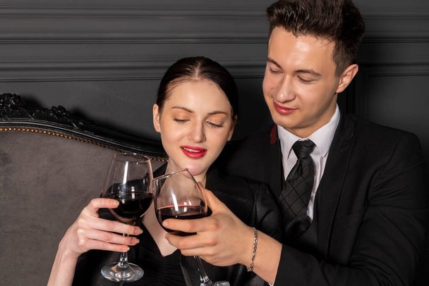 Ein junges Paar, elegant und stilvoll gekleidet, verbringt eine romantische Zeit mit Weingläsern in der Hand auf einer Vintage-Couch in einem wunderschönen dunklen Raum im gotischen Stil. Ein romantisches Date. - Foto, Bild