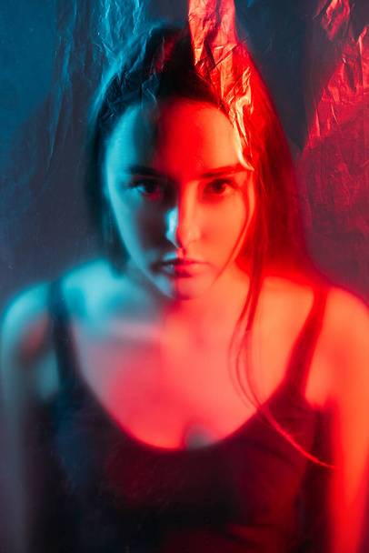Bipolar disorder. Split personality. Mental crisis. Red blue color light defocused art portrait of serious woman face behind distressed transparent polyethylene plastic film wall. - Φωτογραφία, εικόνα