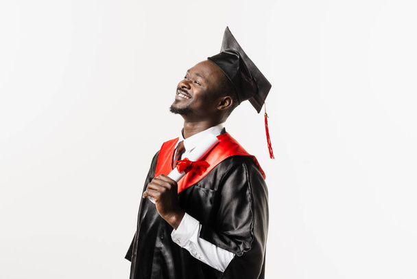 Absolvent afrického muže s magisterským titulem v černých maturitních šatech a čepicí na bílém pozadí. Šťastný africký student úspěšně absolvoval univerzitu s vyznamenáním - Fotografie, Obrázek
