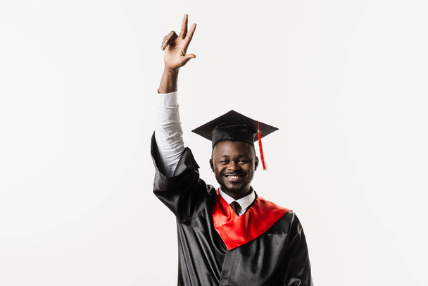 Happy african student in black graduation gown and cap raises his hands above head on white background. Graduate african man is graduating college and celebrating academic achievement - Photo, Image