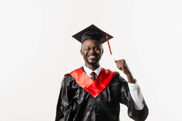 Graduate african man is graduating college and celebrating academic achievement. Happy african student in black graduation gown and cap raises his hands above head on white background - Photo, Image