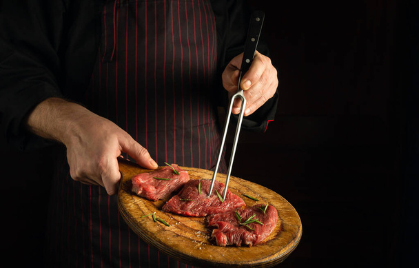Szef kuchni trzyma widelec w ręce i deskę do krojenia ze stekami wołowymi przed grillem. Ciemne miejsce na recepturę restauracji lub menu hotelowe - Zdjęcie, obraz