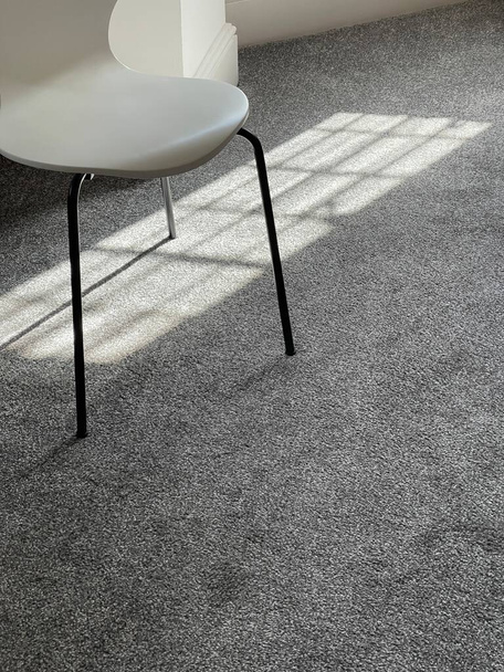 Schnappschuss eines weißen modernen Stuhls auf grauem Teppich, Sonne scheint durch das Fenster in die Dachgeschosswohnung - Foto, Bild
