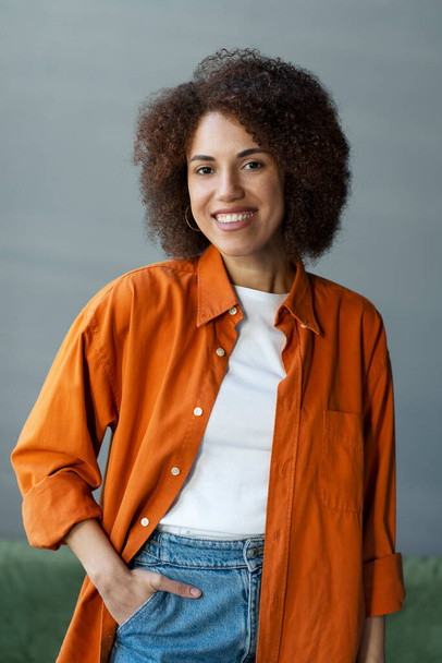Gülümseyen, kendine güvenen Afro-Amerikan bir kadının portresi şık, turuncu bir tişört giymiş, evde dikiliyor. Başarılı kıvırcık saçlı üniversite öğrencisi kamera ve eğitim konseptine bakıyor. - Fotoğraf, Görsel