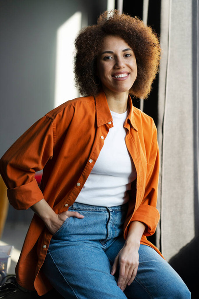 Аутентичный портрет красивой улыбающейся афроамериканки в стильной оранжевой футболке, джинсах, сидящих дома. Счастливая уверенная кудрявая хипстерша смотрит в камеру - Фото, изображение