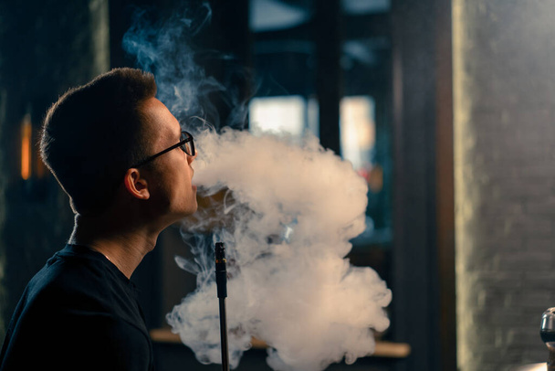 Um hookah homem de óculos fuma um cachimbo de narguilé tradicional Um homem exala fumaça em café narguilé ou lounge bar close-up - Foto, Imagem
