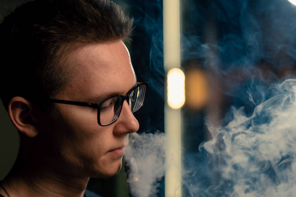 Ένας άντρας με γυαλιά καπνίζει μια παραδοσιακή πίπα, ένας άντρας εκπνέει καπνό σε ένα καφέ ή μπαρ κοντά. - Φωτογραφία, εικόνα