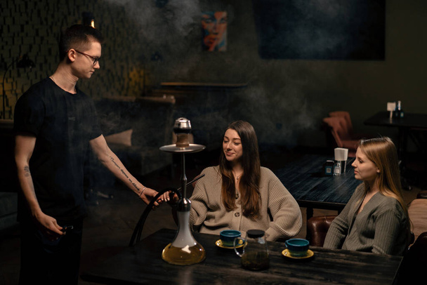 young girlfriends chat in a lounge bar and a hookah man brings hookah and offers to try it - Photo, Image
