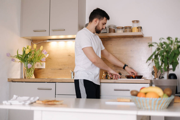 Komea mies valmistamassa ruokaa kotona. Nuori mies yllättää rakkaan naisen ja tekee aamiaista keittiössä. Laadukas kuva - Valokuva, kuva