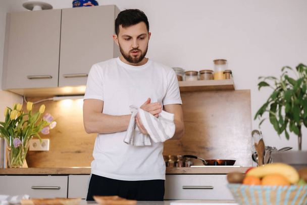 Man wipes hands with a white towel. Handsome bearded man preparing breakfast on kitchen. High quality photo - Foto, Imagen