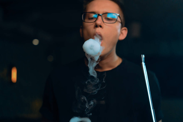 A man in glasses smokes a traditional hookah pipe A man exhales rings of thick smoke in hookah cafe or lounge bar - Photo, Image