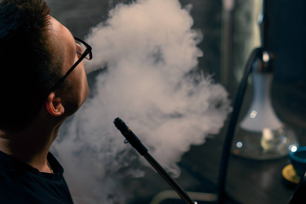 Muž v brýlích kouří tradiční dýmku z hadice Muž vydechuje hustý kouř v kavárně hookah nebo v baru - Fotografie, Obrázek