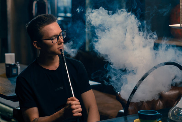 Un hombre con gafas fuma una pipa tradicional de narguile Un hombre exhala humo espeso en la cafetería de narguile o en el bar salón - Foto, imagen