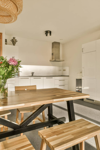 a dining table and bench in a kitchen with white cupboards behind it, there is a vase of flowers on the table - Fotoğraf, Görsel
