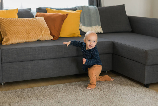 Toddler boy laughing having fun standing near sofa in living room at home. Adorable baby making first steps alone. Happy childhood and child care - Fotó, kép