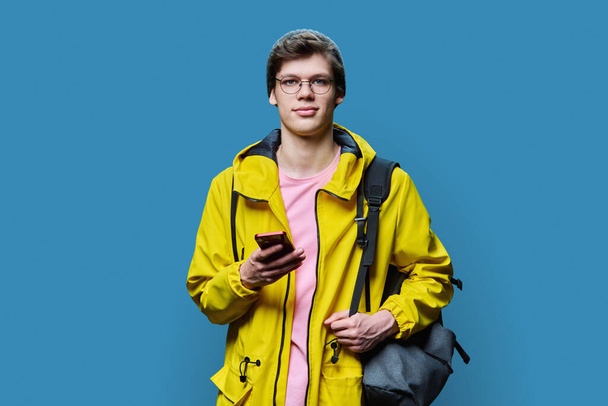 Trendy young male in yellow jacket hat using smartphone on blue studio background. Handsome hipster guy with backpack looking at camera - Valokuva, kuva