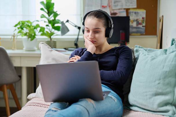 Teen girl in headphones looking at laptop screen, lying on couch at home, leisure relaxation learning communication. Serious attentive teenage female watching video, internet online technology concept - Фото, изображение