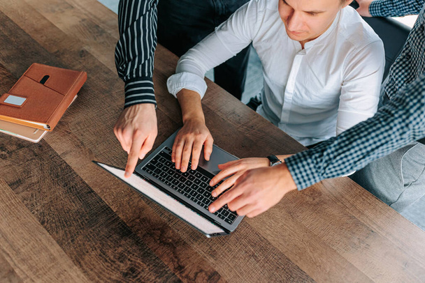 Bovenaanzicht van een mans handen werkend op een laptop in een modern kantoor met een bruisend personeelsbestand zichtbaar op de achtergrond. close-up van een mans handen op een laptop, met andere handen bereiken - Foto, afbeelding
