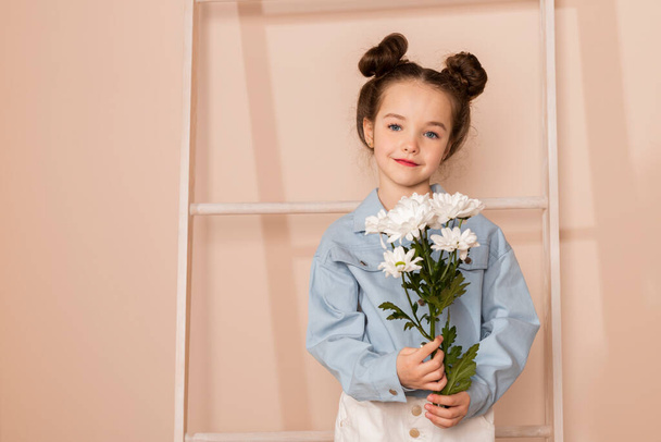 Portrait of a charming little girl, well dressed in a blue shirt and white pants or trousers and with a slight makeup and double bun hair. Isolated on pastel beige background. - Fotó, kép