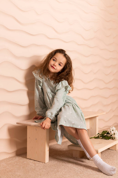 Portrait d'une charmante petite fille, bien habillée dans une robe vert pastel avec un léger maquillage et les cheveux bruns lâches posant avec des fleurs sur un court banc contre un mur beige ondulé. - Photo, image