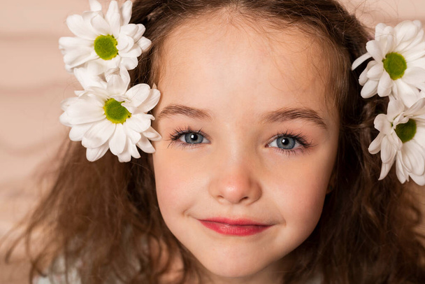 Κοντινό πλάνο Πορτρέτο ενός γοητευτικού μικρού κοριτσιού, καλοντυμένου με παστέλ πράσινο φόρεμα με ελαφρύ μακιγιάζ και καστανά χαλαρά μαλλιά, ποζάροντας με χαμομήλι πάνω σε μπεζ κυματιστό φόντο. - Φωτογραφία, εικόνα