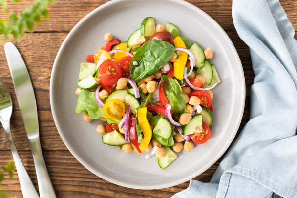 Vegetarian vegetable salad with chickpeas, tomatoes, red onions, cucumbers and lettuce. Vegetarian food rich in protein and fiber. Vegetable salad on wooden table top view.  - Photo, Image