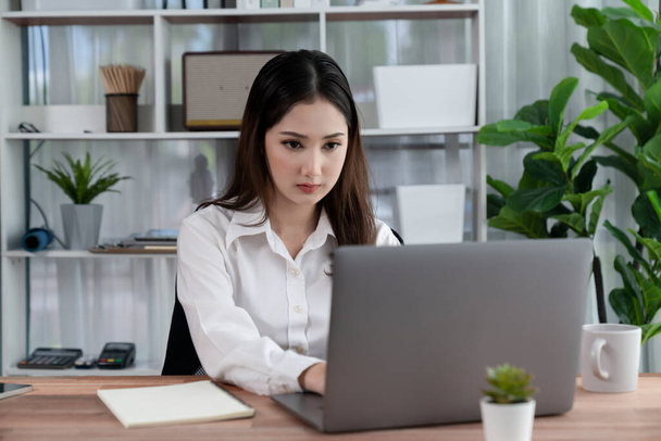 Młoda azjatycka entuzjastyczna bizneswoman w nowoczesnym biurze za pomocą laptopa do pracy z filiżanką kawy. Staranna i atrakcyjna kobieta biurowa pracująca nad notebookiem komputerowym w swojej przestrzeni biurowej. - Zdjęcie, obraz
