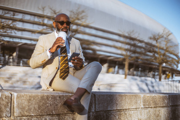 A bald man with a full white-bearded face holding a phone and coffee, dressed in a cream suit with a yellow striped tie, sitting on stone steps wearing black sunglasses - Photo, Image