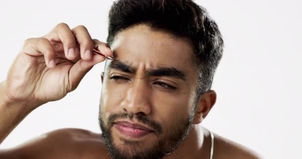 Man grooming his eyebrows with a tweezer in studio for self care, beauty and cleanliness. Hair removal, tweezing and male model from India doing facial epilation plucking routine by white background - Footage, Video