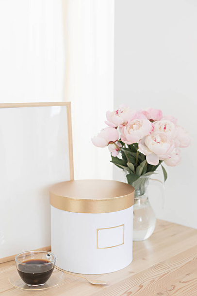 Vertical frame and gift box mockup on a wooden table in the kitchen. Glass jug with a bouquet of pink peonies and a cup of black coffee. Scandinavian style interior - Photo, image