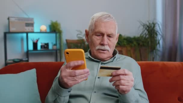 Dědeček senior muž pomocí kreditní karty a smartphone při převodu peněz, nakupuje on-line nakupování bezhotovostní, objednávat doručení jídla, platit účty doma byt uvnitř. Starší muž na gauči - Záběry, video