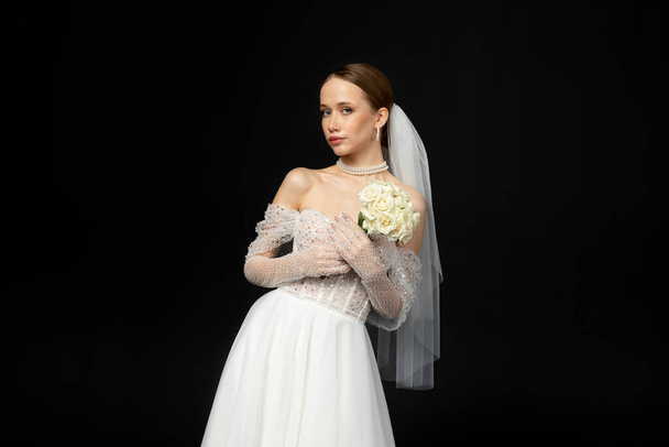 Een mooi charmant meisje, de bruid in een luxe en elegante witte trouwjurk, poseert teder met een sierlijk boeket witte rozen. Geïsoleerd op een zwarte achtergrond. - Foto, afbeelding