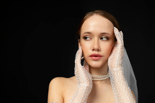Close-up Portret van een mooi charmant meisje, de bruid in een luxe en elegante witte trouwjurk, poseert en gebaart teder. Geïsoleerd op zwarte achtergrond. handen die het gezicht inlijsten. - Foto, afbeelding