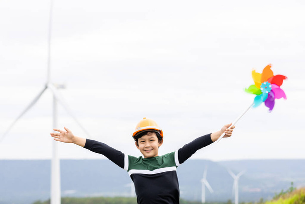Progressive young asian boy playing with wind pinwheel toy in the wind turbine farm, green field over the hill. Green energy from renewable electric wind generator. Windmill in the countryside concept - Valokuva, kuva