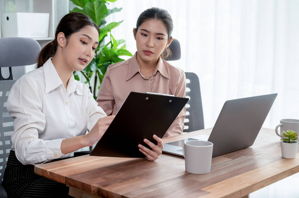 Two young office lady colleagues collaborating in modern office workspace, engaging in discussion and working together on laptop, showcasing their professionalism as modern office worker. Enthusiastic - 写真・画像
