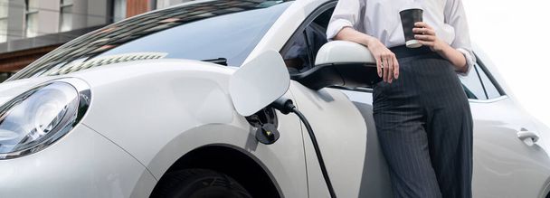 Closeup progressive suit-clad businesswoman with her electric vehicle recharge her car on public charging station in modern city with power cable plug and renewable energy-powered electric vehicle. - Foto, imagen