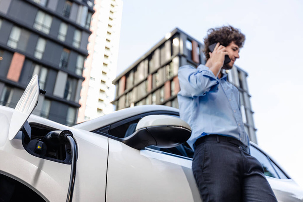 Progressive businessman talking on the phone, leaning on electric car recharging with public EV charging station, apartment condo residential building on the background as green city lifestyle. - 写真・画像
