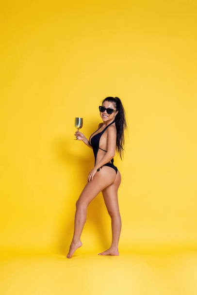 Hermosa chica asiática en un traje de baño negro y gafas de sol sobre un fondo amarillo. foto de alta calidad - Foto, imagen