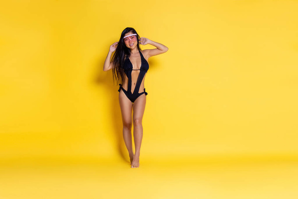  Beautiful asian girl in a black swimsuit and sunglasses on a yellow background. high quality photo - Foto, Imagem