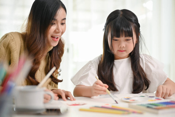 Beautiful and attractive Asian mom helps her little daughter with coloring on paper while having fun family time at home together. - Фото, изображение