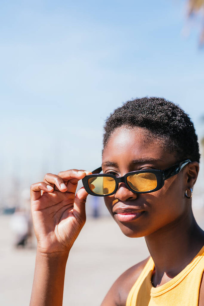pionowy portret atrakcyjnej afrykańskiej kobiety zdejmującej modne żółte okulary przeciwsłoneczne - Zdjęcie, obraz