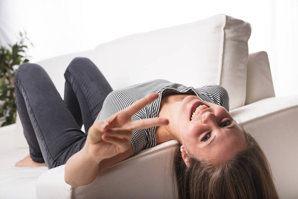 Eine junge Frau liegt auf dem Rücken auf einem weißen Sofa, den Kopf kopfüber auf der Armlehne, barfuß, zeigt ein V für Siegeszeichen und lacht fröhlich. Weiche Haare hängen, verschwommener Pflanzenhintergrund, reichlich Kopie  - Foto, Bild