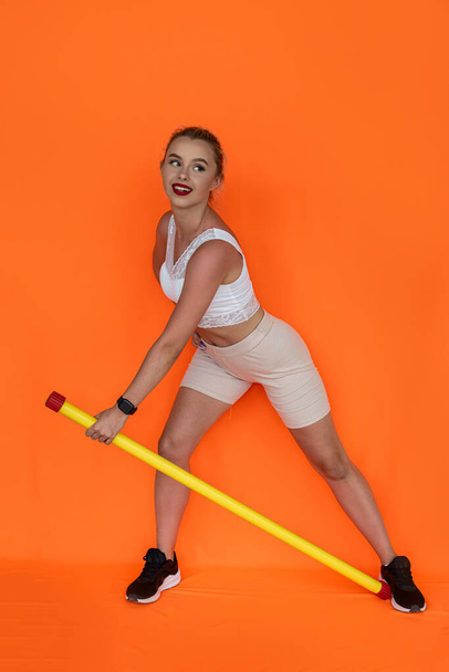 una chica joven con un cuerpo atlético hace un ejercicio de fitness con un palo de fitness en el fondo. Concepto de chica deportiva - Foto, imagen