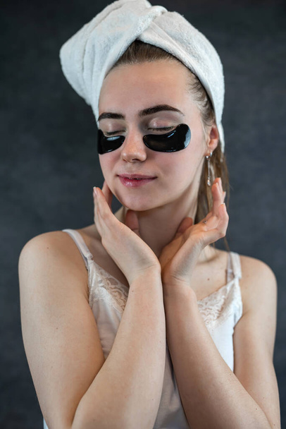 νεαρό χαριτωμένο κορίτσι σε λευκό πετσέτα καθαρό δέρμα με μαύρα μπαλώματα σιλικόνης που απομονώνονται σε μαύρο φόντο. Επιθέματα για τη φροντίδα του δέρματος μαύρους κύκλους κάτω από τα μάτια - Φωτογραφία, εικόνα